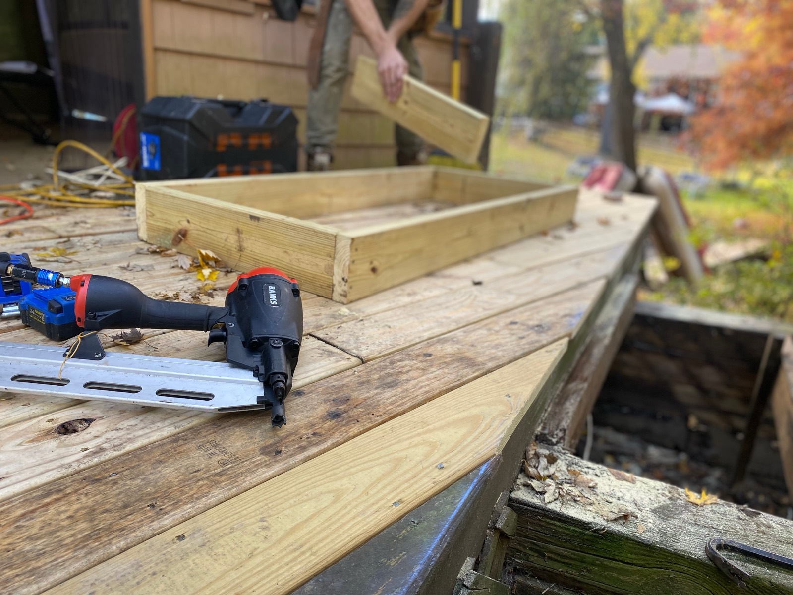 A deck builder carrying a pressure-treated board toward a framed section on a partly rebuilt backyard deck, with a framing nailer resting on the new boards