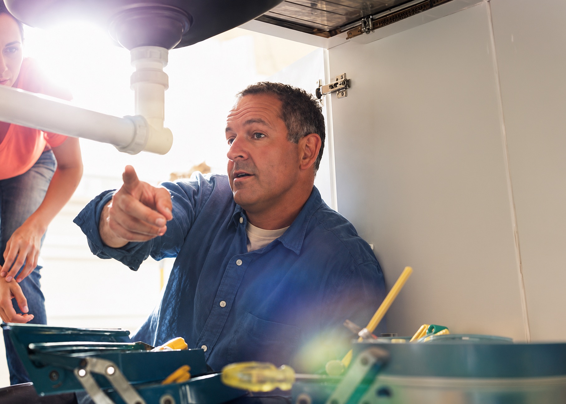 A plumber working under a kitchen sink, pointing out the repair to the homeowner